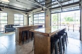 This single-floor commercial space blends industrial design with warm, inviting elements to create a versatile and functional environment. The open-plan layout is anchored by polished concrete floors and soaring ceilings with exposed ductwork and black steel beams, lending the space an authentic industrial character. Pendant lights with matte black fixtures hang throughout, providing focused illumination that complements the natural light pouring in through oversized steel-framed windows and full-height glass garage doors. These doors open to an outdoor patio area, seamlessly connecting the interior to the exterior and offering views of the surrounding greenery.

The main area features substantial dark wood communal tables with bar-height seating, ideal for collaborative work or social gatherings. A lounge nook is furnished with a leather sofa, a colorful area rug, and a small coffee table, creating a comfortable and casual retreat within the open space.

A dedicated kitchenette area is outfitted with dark wood cabinetry, granite countertops, a stainless steel refrigerator, and a double sink — accented by decorative round mirrors and warm wall sconces. A large vintage-style wall clock and tasteful floral arrangements add personality to the space. A water fountain station is also conveniently integrated near the kitchenette, underscoring the thoughtful, functional design throughout.