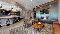 The Club Room at 300 Herb Hill Road in Glen Cove, New York, is a thoughtfully designed communal space that blends contemporary aesthetics with comfortable, functional amenities. The single-floor layout is organized into several distinct yet connected areas, each contributing to an overall atmosphere of modern elegance.

The lounge area serves as a central gathering point, anchored by a sleek built-in fireplace flanked by a mounted flat-screen television and a striking decorative lattice panel. Seating options include a cognac leather sofa, upholstered armchairs, and mid-century inspired accent tables, all set atop a patterned area rug over light wood-style flooring. Exposed beam detailing at the ceiling adds architectural character to the space.

Adjacent to the lounge, a well-appointed wet bar features glossy dark tile backsplash, illuminated open shelving, a white quartz countertop, and stainless steel appliances, making it well-suited for entertaining. A bar counter with decorative pendant lighting and black metal stools provides additional seating.

The dining area accommodates a large wooden table surrounded by tufted upholstered chairs, bathed in natural light from expansive windows and sliding glass doors. These doors open directly onto an outdoor terrace furnished for al fresco gatherings. Throughout, textured stone-look wall cladding, gold pendant accents, and generous glazing create a cohesive, upscale ambiance.