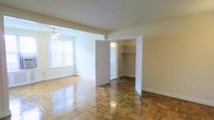 This single-floor space showcases a timeless interior defined by its distinctive parquet hardwood flooring, which features an intricate geometric pattern finished to a high gloss. The layout is open and airy, with soft neutral-toned walls that complement the warm wood tones underfoot. Natural light filters in through a large window dressed with vertical blinds, illuminating the space and enhancing its welcoming ambiance. A ceiling fan is centrally positioned to provide year-round comfort. A notable feature of the space is the generously sized walk-in closet, accessible through double doors, offering built-in shelving and a hanging rod — providing ample storage. An additional door leads to a connected room, suggesting a well-considered flow between spaces. White baseboards and door trim provide clean architectural contrast throughout. The overall character of the space is classic and understated, offering a solid foundation that suits a variety of uses. The combination of functional storage, natural light, and traditional flooring details gives the space a practical yet characterful quality.