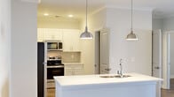 Located at 1025 Hirschfield Dr in Fayetteville, NC, this single-floor residence offers a thoughtfully designed open-concept layout that seamlessly connects the living and kitchen areas. The kitchen is a standout feature, showcasing white shaker cabinetry, a large center island with a double sink, sleek white countertops, stainless steel appliances including a refrigerator, range, microwave, and dishwasher, and elegant pendant lighting. A dedicated pantry with wire shelving provides generous storage. Crown molding and hardwood-style flooring flow throughout the residence, lending a clean and cohesive aesthetic. The living area is bright and spacious, with ceiling fans, crown molding, and glass doors that open onto a private balcony. The balcony features a warm wood-paneled ceiling, classic railing, and open views of the surrounding landscape and mature trees. Bedrooms are comfortably sized, filled with natural light through slatted window blinds, and include walk-in closets with multi-tier wire shelving systems. Bathrooms are finished with white cabinetry, combination tub-shower units, and hardwood-style flooring that complements the rest of the home. The overall ambiance is bright, modern, and move-in ready.