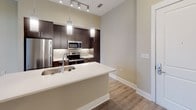 This single-floor residence offers a thoughtfully designed open-concept layout that seamlessly connects the kitchen, dining, and living areas. Warm-toned hardwood-style flooring flows throughout the space, complementing the neutral beige and cream wall palette that creates a bright, welcoming atmosphere.

The kitchen is a standout feature, anchored by a large white-topped island with an integrated sink and pendant lighting overhead. Rich dark cabinetry lines the walls, paired with light quartz countertops, a tile backsplash, and a full suite of stainless steel appliances including a refrigerator, range, and microwave. Track lighting illuminates the cooking area with precision.

The open living area benefits from soaring ceilings and generous natural light, courtesy of oversized French doors that lead to an outdoor space. A ceiling fan adds both function and style to this airy room.

The bedroom, accessed via a sliding barn door, offers a cozy and private retreat. The bathroom is finished with clean, modern fixtures, including a dark-framed vanity mirror, undermount sink set in a light quartz countertop, dark wood cabinetry below, and polished chrome accessories. A barn-style sliding door adds an architectural accent to the transition between spaces, tying together the home's contemporary design aesthetic.