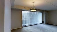 This single-floor space features a spacious living area defined by its clean, understated design. The room's most prominent feature is a wall of floor-to-ceiling sliding glass doors dressed with vertical blinds, allowing an abundance of natural light to filter in while offering views of the surrounding urban landscape. The exterior view reveals a neighboring building and open sky, situating this space within an urban setting.

The living area is finished with neutral-toned carpet, contributing to a warm and comfortable atmosphere. A classic chain-hung pendant light with a bowl-shaped shade serves as the central lighting fixture, adding a modest decorative touch to the otherwise simple ceiling. The walls are painted in a soft, neutral tone that complements the room's muted color palette.

HVAC vents are integrated into both the ceiling and baseboard area near the glass doors, ensuring climate control throughout the space. The overall design is functional and straightforward, with an emphasis on openness and natural light. The layout is well-suited for a variety of furniture arrangements, making it a versatile and practical living environment.