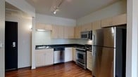 This single-floor space showcases a clean, contemporary design with thoughtful finishes throughout. The kitchen is a standout feature, offering a functional and stylish layout equipped with a full suite of stainless steel appliances, including a gas range, built-in microwave, dishwasher, and a full-size refrigerator. Light-toned cabinetry extends to the ceiling, providing generous storage space, while dark countertops create a striking contrast that adds depth and sophistication to the room. A stainless steel sink with under-cabinet lighting completes the workspace. The rich, dark hardwood flooring runs throughout, adding warmth and a sense of continuity to the space. Track lighting mounted on the ceiling provides adjustable, focused illumination, enhancing both functionality and ambiance. The overall design palette — combining light cabinetry, dark surfaces, and warm wood tones — creates a balanced, modern aesthetic. The layout is efficient and well-organized, making excellent use of the available space while maintaining an open and inviting feel. This is a well-finished, move-in-ready space suited for those who appreciate modern design and practical living.