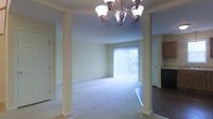 This two-floor residence is thoughtfully designed to balance elegance and functionality. The main living area serves as the centerpiece of the home, characterized by soft, neutral-toned walls and plush wall-to-wall carpeting that creates a warm and comfortable atmosphere. Decorative columns frame the space, adding architectural interest and a sense of structure without enclosing the open layout.

A standout feature of the living area is the elegant chandelier overhead, fitted with multiple light fixtures that provide warm, ambient illumination. The room is further enhanced by large sliding glass doors fitted with vertical blinds, which allow abundant natural light to pour in while offering access to an outdoor area beyond. The greenery visible through the doors adds a pleasant natural backdrop.

Adjacent to the living area, a transition to darker hardwood-style flooring signals a shift toward a dining or kitchen space, suggesting a natural flow between living and entertaining zones. The overall design reflects a clean, classic aesthetic with practical finishes suited to everyday living across both floors.