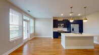Located at 100 Fulton St SE in Atlanta, GA, this single-floor unit showcases a thoughtfully designed open-concept layout that seamlessly connects the kitchen and living areas. The kitchen is a standout feature, outfitted with bold navy blue cabinetry accented by gold hardware, stainless steel appliances including a French door refrigerator, electric range, and built-in microwave. A large central island with a crisp white quartz countertop and matte black sink serves as the focal point of the space, illuminated by a pair of elegant brass globe pendant lights. Warm-toned hardwood flooring flows throughout the main living areas, enhancing the brightness brought in by multiple large windows dressed with horizontal blinds. A glass door provides direct access to an outdoor area, offering views of the Atlanta skyline. The bedroom features plush carpeting, a ceiling fan, built-in shelving, and a generous window framing city views. The bathroom is complemented by an adjoining walk-in closet with wire shelving. A stacked washer and dryer unit is conveniently integrated into the layout, adding practical functionality. The overall design balances modern aesthetics with comfortable, everyday livability.