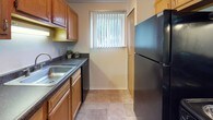 This single-floor residential unit offers a practical and comfortable layout designed for everyday living. The kitchen is efficiently arranged with warm wood cabinetry, dark countertops, a stainless steel sink, built-in dishwasher, and a full-size black refrigerator. A window fitted with vertical blinds allows natural light to fill the space, creating a pleasant cooking environment with tile flooring underfoot.

The living area is generously sized, featuring neutral-toned carpet, a front entry door, and a large window with vertical blinds that invites ample natural light. A wall-mounted air conditioning unit provides climate control, and the open layout offers flexible furniture arrangement options. An adjacent area, separated by a partial wall, includes a baseboard heater, a closet, and a mix of tile and carpet flooring, making it well-suited as a bedroom or multipurpose room.

A second bedroom features soft carpeting and a bright window that frames views of surrounding greenery, creating a calm and airy atmosphere. The bathroom is clean and well-appointed, featuring a wide vanity with a white countertop, an undermount sink with brushed nickel fixtures, warm wood cabinetry below, and a large mirror with overhead lighting. A shower is accessible nearby, completing the functional layout of this well-maintained residential space.