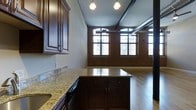 This 2-bedroom, 2-bathroom residence offers a striking blend of industrial character and modern finishes across a single, open floor plan. The space is defined by soaring ceilings adorned with dark wood beams and exposed ductwork, lending an authentic loft aesthetic throughout. Expansive arched windows with multi-pane detailing line the exterior walls, flooding the interior with abundant natural light while showcasing the building's classic brick facade.

The kitchen is a standout feature, boasting rich dark mahogany cabinetry with detailed molding, polished granite countertops, and a central island that provides additional prep space and storage. Pendant lighting above the island adds warm, focused illumination, complementing the open connection to the main living area.

The living space extends generously from the kitchen, featuring warm hardwood flooring that runs throughout and exposed brick accent walls that add texture and depth to the interior. The combination of these raw architectural elements with refined finishes creates an inviting and sophisticated ambiance. This residence is well-suited for those who appreciate urban character paired with comfortable, contemporary living.