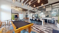 This thoughtfully designed single-floor amenity space offers a generously sized, multi-functional layout that seamlessly blends social, recreational, and relaxation areas. The grand entrance lobby sets an elegant tone with soaring ceilings, powder-blue walls, large arched windows, and a striking circular chandelier overhead. Variegated wood-look tile flooring runs throughout, providing visual continuity across each zone.

The main open area is anchored by dramatic exposed dark wood ceiling beams, from which an array of globe pendant lights descend, casting warm ambient light across the space. A comfortable lounge zone features a large sectional sofa and distinctive hexagonal marble coffee tables paired with vibrant amber leather accent chairs, complemented by a wall-mounted flat-screen TV on a rich wood panel.

Adjacent to the lounge, a game area houses a full-size pool table and connects to a bar-height counter with sleek black seating—ideal for social gatherings. A dedicated dining and café zone provides additional seating alongside a service counter equipped with beverage appliances. Multiple sets of glass French doors open to outdoor views, flooding the interior with natural light and maintaining a bright, airy atmosphere throughout.
