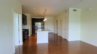 This single-floor space is defined by its open-concept layout, seamlessly connecting the kitchen and living areas across rich, warm-toned hardwood flooring. The kitchen is a standout feature, outfitted with dark espresso cabinetry that extends to the ceiling, providing generous storage. A professional-grade range and stainless steel French door refrigerator with built-in ice and water dispenser anchor the cooking area. A central island with a white base and granite countertop serves as the kitchen's focal point, illuminated by a trio of pendant lights suspended from the ceiling, adding both function and visual interest. Recessed lighting throughout ensures the space remains bright and well-lit. The open floor plan allows for a natural flow between the kitchen and the adjacent living space, which offers ample room for a variety of furniture arrangements. Crisp white walls and white-paneled doors contrast cleanly with the darker finishes of the kitchen, creating a balanced, contemporary aesthetic. The overall ambiance is polished and practical, with quality finishes that lend the space a refined yet livable character.
