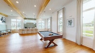 Located at 3795 Bismarck Ct in Wildwood, Florida, this single-story clubhouse offers a well-appointed communal space defined by modern farmhouse architecture and thoughtfully designed interiors. Warm hardwood flooring flows continuously throughout, complemented by exposed natural wood ceiling beams, shiplap detailing, and recessed lighting that collectively create an inviting, airy atmosphere.

The expansive main lounge serves as the heart of the space, featuring a dramatic coffered ceiling with timber beams, a striking floor-to-ceiling stacked stone fireplace, a wall-mounted television, and comfortable seating arrangements. Contemporary chandeliers and pendant fixtures add refined lighting accents throughout.

A dedicated game room accommodates a full-size pool table and foosball table, bathed in natural light from large windows with views of the surrounding landscape. Adjacent to this area, a stylish kitchenette and bar feature a long white island with bar seating, globe pendant lights, sage green subway tile backsplash, under-counter beverage refrigerators, and a coffee station. Built-in banquette seating with channel-tufted upholstery provides a cozy nook nearby.

Tall windows throughout the space flood every area with natural light while offering pleasant outdoor views, enhancing the open, social character of this well-crafted community gathering facility.