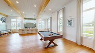 Located at 3795 Bismarck Ct in Wildwood, Florida, this single-story clubhouse offers a well-appointed communal space defined by modern farmhouse architecture and thoughtfully designed interiors. Warm hardwood flooring flows continuously throughout, complemented by exposed natural wood ceiling beams, shiplap detailing, and recessed lighting that collectively create an inviting, airy atmosphere.

The expansive main lounge serves as the heart of the space, featuring a dramatic coffered ceiling with timber beams, a striking floor-to-ceiling stacked stone fireplace, a wall-mounted television, and comfortable seating arrangements. Contemporary chandeliers and pendant fixtures add refined lighting accents throughout.

A dedicated game room accommodates a full-size pool table and foosball table, bathed in natural light from large windows with views of the surrounding landscape. Adjacent to this area, a stylish kitchenette and bar feature a long white island with bar seating, globe pendant lights, sage green subway tile backsplash, under-counter beverage refrigerators, and a coffee station. Built-in banquette seating with channel-tufted upholstery provides a cozy nook nearby.

Tall windows throughout the space flood every area with natural light while offering pleasant outdoor views, enhancing the open, social character of this well-crafted community gathering facility.