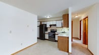 This single-floor space presents a practical and well-organized layout, with the kitchen serving as a central feature. The kitchen is equipped with stainless steel appliances, including a refrigerator and an electric range with a ventilation hood above. Cabinetry combines warm medium-toned wood lower cabinets and a tall pantry-style unit with white upper cabinets, offering ample storage throughout. A light-colored countertop runs along the workspace, with a single-basin sink positioned beneath under-cabinet lighting for added functionality. The backsplash behind the range provides a subtle contrast to the overall neutral palette. The kitchen floor features light-toned vinyl or tile, transitioning into carpeted flooring visible in the adjacent area, suggesting an open flow between the kitchen and a neighboring living space. White walls and a textured ceiling reflect the overhead flush-mount lighting, keeping the interior bright and clean. Wood-toned baseboards throughout add a cohesive finishing detail. The layout is straightforward and efficient, making good use of the available space while maintaining a comfortable, livable atmosphere.