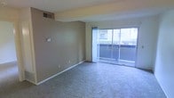 This single-story space features an open living area finished with wall-to-wall carpeting in a neutral gray tone, providing a comfortable and understated foundation throughout. The walls are painted in a warm taupe, complementing the overall subdued color palette of the interior. A ceiling-mounted HVAC vent ensures climate control, while a wall-mounted thermostat offers convenient temperature regulation.

The most prominent feature of the living area is the full-width sliding glass door, framed by sheer white curtains that allow natural light to filter softly into the room. The slider opens directly to an outdoor patio space, offering views of manicured greenery and adjacent structures, creating a pleasant visual connection between the interior and the outdoors. The patio extends the functional living space and provides an accessible outdoor retreat.

White baseboards trim the perimeter of the room, adding a clean contrast to the carpeted flooring and taupe walls. The space is straightforward in layout, offering an open, flexible floor plan well-suited for a variety of furniture arrangements.