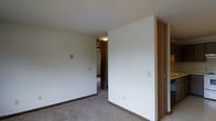 This single-floor space presents a straightforward and functional interior design. The image reveals a carpeted room with neutral white walls and dark wood baseboards that provide a subtle contrast to the light-colored flooring. A sliding closet with warm wood-toned doors is a notable feature, offering built-in storage directly within the room.

The space transitions into an adjoining area visible through an open doorway, where additional wood-finish cabinetry or closet units can be seen, suggesting consistent storage solutions throughout. A flush-mount ceiling light fixture provides overhead illumination in the connecting space.

The walls are clean and unadorned, contributing to a minimalist aesthetic throughout. Electrical outlets and light switch plates are positioned at standard heights along the walls. The carpet extends throughout the visible areas, lending a uniform, cohesive feel to the interior.

Overall, the space is characterized by its simple, practical design with consistent use of neutral tones, wood accents on the closet doors and trim, and wall-to-wall carpeting that creates a quiet, understated atmosphere suitable for everyday living.