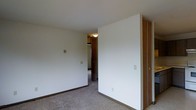 This single-floor space presents a straightforward and functional interior design. The image reveals a carpeted room with neutral white walls and dark wood baseboards that provide a subtle contrast to the light-colored flooring. A sliding closet with warm wood-toned doors is a notable feature, offering built-in storage directly within the room.

The space transitions into an adjoining area visible through an open doorway, where additional wood-finish cabinetry or closet units can be seen, suggesting consistent storage solutions throughout. A flush-mount ceiling light fixture provides overhead illumination in the connecting space.

The walls are clean and unadorned, contributing to a minimalist aesthetic throughout. Electrical outlets and light switch plates are positioned at standard heights along the walls. The carpet extends throughout the visible areas, lending a uniform, cohesive feel to the interior.

Overall, the space is characterized by its simple, practical design with consistent use of neutral tones, wood accents on the closet doors and trim, and wall-to-wall carpeting that creates a quiet, understated atmosphere suitable for everyday living.
