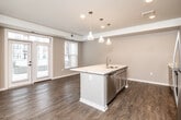 This single-floor unit offers three bedrooms and one bathroom within a thoughtfully designed layout. The open-concept kitchen and living area serves as the heart of the space, featuring rich hardwood-style flooring throughout. The kitchen is equipped with stainless steel appliances, including a refrigerator, range, and microwave, complemented by taupe cabinetry, a subway tile backsplash, and a large central island topped with a marble-look surface. Three decorative pendant lights hang above the island, adding a contemporary touch. The living area benefits from natural light through a glass-panel exterior door flanked by windows, offering views to the outdoors.

The three bedrooms feature neutral carpet flooring and warm-toned walls, providing a calm, functional atmosphere. The primary bedroom includes a walk-in closet with wire shelving. The shared bathroom features a vanity with under-sink storage and a glass-enclosed shower. A dedicated in-unit laundry closet houses a full-size washer and dryer. A connecting hallway with hardwood-style flooring ties all rooms together, creating a cohesive and efficient flow throughout the unit.