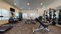 This fitness center is designed to accommodate a comprehensive range of workout styles within a single, open-plan space. The room features neutral white walls, recessed lighting, and durable patterned carpet flooring throughout, creating a clean and functional environment. Multiple windows along the perimeter allow natural light to fill the space, contributing to an open and energizing atmosphere.

The cardio zone is well-stocked with treadmills, an elliptical trainer, a stationary bike, and a rowing machine, with wall-mounted flat-screen televisions positioned for viewing during exercise. A dedicated strength training area includes a power rack loaded with Olympic weight plates, an adjustable Matrix bench, and a full dumbbell rack with a broad range of weights. A cable machine provides additional resistance training options. A large wall mirror in the strength zone enhances the sense of space and allows users to monitor form.

Functional training accessories, including stability balls, resistance bands, and medicine balls, are neatly organized along one wall. The layout provides generous floor space between equipment, allowing for comfortable movement and safe use throughout the facility.
