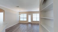This single-floor space offers a clean, modern aesthetic defined by warm neutral tones and an open, airy layout. The main living area features smooth hardwood-style flooring in a warm gray-brown finish, complementing the crisp white ceilings and soft beige walls throughout. Crown molding adds a subtle architectural detail, lending a polished, finished look to the space.

Natural light is a standout feature, provided by multiple large windows dressed with horizontal blinds, flanking a central exterior door that reinforces the bright, welcoming atmosphere. A pendant light fixture hangs centrally, offering both functional and decorative illumination.

To one side of the room, built-in open shelving provides practical storage and display space, seamlessly integrated into the wall for a clean, uncluttered appearance. The overall layout is versatile and well-proportioned, offering ample room for comfortable furnishing arrangements.

The space presents a move-in-ready condition with a neutral palette that accommodates a wide range of interior styles. Its combination of natural light, quality finishes, and thoughtful built-in features makes it a functional and visually appealing living environment.
