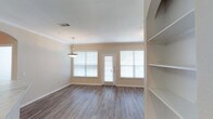 This single-floor space offers a clean, modern aesthetic defined by warm neutral tones and an open, airy layout. The main living area features smooth hardwood-style flooring in a warm gray-brown finish, complementing the crisp white ceilings and soft beige walls throughout. Crown molding adds a subtle architectural detail, lending a polished, finished look to the space.

Natural light is a standout feature, provided by multiple large windows dressed with horizontal blinds, flanking a central exterior door that reinforces the bright, welcoming atmosphere. A pendant light fixture hangs centrally, offering both functional and decorative illumination.

To one side of the room, built-in open shelving provides practical storage and display space, seamlessly integrated into the wall for a clean, uncluttered appearance. The overall layout is versatile and well-proportioned, offering ample room for comfortable furnishing arrangements.

The space presents a move-in-ready condition with a neutral palette that accommodates a wide range of interior styles. Its combination of natural light, quality finishes, and thoughtful built-in features makes it a functional and visually appealing living environment.
