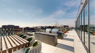 This rooftop terrace is a thoughtfully designed outdoor amenity space elevated above the surrounding cityscape, offering sweeping panoramic views of the urban skyline in every direction. The layout is generously proportioned, wrapping in an L-shaped configuration that accommodates a diverse array of functional zones across its open-air footprint.

At the heart of the terrace sits a sleek swimming pool, framed by bold red and navy sun loungers arranged along its perimeter. A modern pergola structure overhead provides partial shade to an adjacent lounge area furnished with plush, oversized seating and accent tables, enhanced by café string lighting for evening ambiance. An outdoor television mounted to the building's exterior wall makes this a versatile entertainment zone.

The outdoor kitchen area features multiple stainless steel grills set into built-in countertop stations, offering ample space for al fresco cooking and dining. Nearby, a dedicated lounge zone centers around a concrete fire pit table surrounded by teal and red accent chairs and cylindrical ottomans, complemented by planters with seasonal greenery throughout. Glass railings along the perimeter preserve the expansive city views while maintaining an open, airy atmosphere across the entire space.