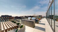 This rooftop terrace is a thoughtfully designed outdoor amenity space elevated above the surrounding cityscape, offering sweeping panoramic views of the urban skyline in every direction. The layout is generously proportioned, wrapping in an L-shaped configuration that accommodates a diverse array of functional zones across its open-air footprint.

At the heart of the terrace sits a sleek swimming pool, framed by bold red and navy sun loungers arranged along its perimeter. A modern pergola structure overhead provides partial shade to an adjacent lounge area furnished with plush, oversized seating and accent tables, enhanced by café string lighting for evening ambiance. An outdoor television mounted to the building's exterior wall makes this a versatile entertainment zone.

The outdoor kitchen area features multiple stainless steel grills set into built-in countertop stations, offering ample space for al fresco cooking and dining. Nearby, a dedicated lounge zone centers around a concrete fire pit table surrounded by teal and red accent chairs and cylindrical ottomans, complemented by planters with seasonal greenery throughout. Glass railings along the perimeter preserve the expansive city views while maintaining an open, airy atmosphere across the entire space.