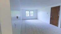 This single-floor space presents a clean and minimalist interior defined by its bright white walls and soft wall-to-wall carpeting in a neutral tone. The open room is filled with natural light, which streams in through a pair of double-hung windows positioned along the far wall, offering views of surrounding greenery. Below the windows, a baseboard heating unit runs the length of the wall, providing an efficient heating solution integrated seamlessly into the room's design. The overall palette is light and airy, contributing to a sense of openness throughout the space. The walls are smooth and freshly maintained, complementing the understated aesthetic. A door is visible along one side, suggesting straightforward access and a practical layout. The space is vacant and move-in ready, allowing for flexible furnishing arrangements to suit a variety of needs. With its uncomplicated design, neutral finishes, and ample natural light, this space offers a calm and functional environment well-suited for comfortable everyday living.