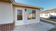This single-story residential unit offers a straightforward and functional layout designed for comfortable everyday living. The exterior features light vinyl siding, a private entry door with an adjacent wall-mount lantern fixture, and a large sliding glass window, all accessible via a concrete patio — providing a welcoming approach to the unit.

Inside, the main living area is generously proportioned, finished with neutral-toned patterned carpeting and warm wood baseboards that run throughout. A ceiling fan with a light fixture is centrally positioned, offering both airflow and ambient lighting. Natural light enters through a large sliding glass door, which offers views of the surrounding neighborhood and yard area.

The entry foyer provides a smooth transition into the living space, with a full-size exterior door, a sidelight window, and a floor vent indicating a forced-air heating system. Adjacent to the main living area, an open passage leads to a closet and connects to what appears to be a kitchen space with dark cabinetry visible in the background. The walls throughout are finished in a clean, neutral white, creating a bright and versatile interior ready for personalization. Overall, the unit presents a clean, move-in ready condition with a practical and cohesive design.