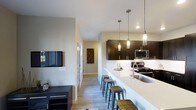 This single-floor space showcases a thoughtfully designed open-concept layout that seamlessly connects the kitchen and living areas. The kitchen is a standout feature, anchored by an expansive white quartz countertop that doubles as a breakfast bar, accommodating multiple industrial-style metal stools for casual dining or entertaining. Dark espresso cabinetry provides a striking contrast to the light countertops and white tile backsplash, while stainless steel appliances, including a gas range and built-in microwave, offer a sleek, contemporary feel. Three elegant glass pendant lights hang above the bar, adding warmth and visual interest to the space. The living area is finished with light hardwood-style flooring that runs throughout, contributing to a cohesive and airy atmosphere. Neutral wall tones keep the space bright and versatile. A wall-mounted flat-screen television is positioned opposite a low-profile media console, completing the living area. A hallway extends toward additional rooms, with decorative wall art adding a personalized touch. Natural light filters through windows, enhancing the open and welcoming ambiance of this modern, functional space.