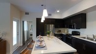 This single-floor space showcases a contemporary open-concept kitchen and dining area designed with a bold contrast of dark and light elements. The kitchen is outfitted with rich, dark espresso cabinetry that extends to the upper walls, providing generous storage while creating a sleek, dramatic aesthetic. Stainless steel appliances, including a refrigerator and gas range with matching microwave, complement the modern design and offer full functionality.

The centerpiece of the space is a large white quartz kitchen island that doubles as a dining surface, comfortably seating multiple guests. Its crisp, bright finish provides a striking contrast against the dark cabinetry. A trio of modern conical pendant lights hang directly above the island, delivering focused task lighting while serving as a standout design feature.

The flooring throughout is hardwood in a warm, medium tone, adding depth and warmth to the space. Recessed lighting supplements the pendant fixtures, ensuring the kitchen is well-lit throughout. A glimpse through an adjacent doorway reveals a connected bedroom with natural light filtering in through draped windows, suggesting a well-connected, cohesive floor plan overall.