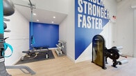 Located at 1320 Via Terrassa in Encinitas, California, this single-floor fitness facility delivers a clean, modern environment designed to inspire performance. The space is anchored by an open-concept workout area with high ceilings supported by exposed white beams, light wood-style flooring, and bright white walls that create an airy, energizing atmosphere. A bold, large-scale graphic mural reading "Harder. Stronger. Faster." in white lettering against a striking blue accent wall serves as a focal motivational centerpiece throughout the space. The facility is well-equipped with a comprehensive range of professional-grade equipment, including a multi-functional cable machine, an Instinct training machine, a rowing ergometer, TRX suspension straps, kettlebells, medicine balls, resistance bands, dumbbells, and a weight bench. A dedicated storage rack keeps accessories neatly organized. A built-in water fountain station provides convenient hydration access, and a wall-mounted mini-split HVAC system ensures year-round climate comfort. A large roll-up garage-style door opens directly to the outdoor parking area, flooding the interior with natural light and offering a seamless indoor-outdoor connection. The surrounding community features mature trees and Spanish-style architecture, contributing to a welcoming and well-maintained environment.