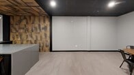 Located at 480 Myrtle St in New Britain, Connecticut, this single-floor commercial space offers a generously sized, open-plan layout ideal for gatherings, events, or collaborative use. The interior is defined by a striking ceiling design that combines dark metal grid framework with warm wood and corrugated metal tile insets, creating a bold industrial-modern aesthetic. Recessed and pendant lighting — including a contemporary globe chandelier — provide versatile illumination throughout the space. Smooth, light-toned large-format tile flooring extends across the entire floor, contrasting elegantly with the dark baseboards and structural columns. The walls are finished in a clean, neutral white, offering a blank canvas that enhances the sense of openness. A textured, vertically ribbed wood-panel accent wall adds warmth and visual depth to one corner of the room. Multiple round wooden tables paired with black metal chairs are arranged throughout, supporting a variety of seating configurations. A doorway leads to what appears to be an adjacent outdoor area, introducing a connection to the exterior. The overall ambiance is contemporary and polished, balancing industrial character with refined, welcoming details.