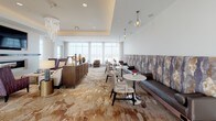 This expansive single-floor space is thoughtfully designed to accommodate a variety of social and functional uses, combining refined aesthetics with practical amenity areas. The layout, as revealed in the floor plan overview, comprises several distinct zones that flow cohesively from one to the next.

The lounge areas are among the most visually striking portions of the space, featuring patterned carpeting with an organic, swirling motif underfoot, floor-to-ceiling sheer drapery filtering abundant natural light, and carefully curated seating arrangements. Upholstered banquettes with bold, abstract fabric backdrops anchor one seating zone, while clusters of armchairs and small tables create intimate conversation areas nearby. Pendant lighting adds warmth overhead, and tall planters with ornamental grasses serve as natural dividers between zones.

The dining area presents herringbone-patterned hardwood flooring, wood-accented tray ceilings, and walls of sheer-curtained windows that flood the room with daylight. A mix of dining tables accommodates both small and larger groups comfortably.

The kitchen is a standout feature, showcasing a warm wood-panel ceiling, a lengthy white quartz island, stainless steel professional-grade appliances, mosaic tile backsplashes, and geometric copper pendant lighting. A dedicated beverage station with built-in refrigeration adds further functionality.

A casual activity nook rounds out the space, featuring a wall-mounted oversized Scrabble board, a television, and modular seating, offering a playful counterpoint to the more formal areas.