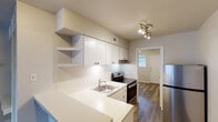 This one-bedroom, one-bathroom unit in Austin, TX offers a thoughtfully designed, single-floor layout that makes excellent use of its compact footprint. The open-concept living and kitchen area is anchored by a peninsula-style countertop that doubles as a breakfast bar, creating a natural division between the two spaces while maintaining an airy, connected feel. The kitchen is well-equipped with white cabinetry, open corner shelving, light-toned countertops, a stainless steel sink, a stainless steel refrigerator, and a stainless steel electric range with a vent hood. A built-in shelving and desk unit adjacent to the kitchen provides convenient storage and a dedicated workspace, adding functional versatility to the living area. Rich hardwood-style flooring runs throughout the main living spaces, adding warmth and cohesion to the neutral color palette. The bedroom features a ceiling fan with light, a window with blinds that allows natural light to filter in, and the same hardwood-style flooring. The bathroom includes a full tub and shower combination with clean white subway tile surround, a toilet, and an overhead cabinet for storage. The overall design is clean, modern, and functional — well-suited for those seeking a streamlined living space in the heart of Austin.