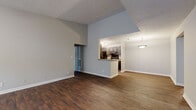This single-floor residence offers a well-designed layout that balances open communal spaces with private living quarters. The living area is immediately impressive, featuring soaring vaulted ceilings with a textured finish and rich, dark wood-style flooring that runs throughout the main areas. Natural light filters in through windows at the entry, creating a bright and welcoming atmosphere. The space flows seamlessly from the living area toward the kitchen, which is accessible via a half-wall peninsula that doubles as additional counter space. The galley-style kitchen is well-appointed with dark gray shaker cabinetry, granite-style countertops, black appliances including a refrigerator, range, and dishwasher, and track lighting overhead. A dedicated laundry or utility closet is accessible directly from the kitchen corridor. The bedrooms are carpeted for comfort and feature neutral gray walls, ample space, and walk-in closets with wire shelving. The bathroom includes a vanity with dark cabinetry, a granite-style countertop, a large mirror, and a combined tub and shower. Throughout the residence, consistent neutral tones, clean white trim, and practical finishes create a cohesive and functional living environment.