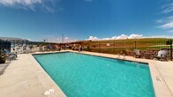 Located at 7175 Dunsmore Rd in Rapid City, South Dakota, this outdoor pool area offers a well-appointed recreational space designed for relaxation and leisure. The centerpiece is a generously sized rectangular swimming pool with clear, blue water, accessible via pool-side entry steps and ladder access points. The pool is framed by a wide, paved sun deck that provides ample room for movement and social gathering.

Surrounding the pool, numerous slatted lounge chairs are arranged to take full advantage of the South Dakota sunshine, offering comfortable spots for sunbathing and unwinding. Outdoor seating sets, including tables and chairs, are also positioned around the deck, making the space suitable for casual outdoor dining or socializing.

The entire pool area is enclosed by a sturdy black iron perimeter fence, featuring a secure gate entry that ensures both safety and controlled access. Decorative potted plants are placed near the gate, adding a touch of greenery to the hardscape environment.

The surrounding landscape opens up to expansive natural terrain, offering open views of rolling hills and wide skies characteristic of the Rapid City region. The building adjacent to the pool features a combination of blue siding and brick accents beneath a pitched shingle roof, complementing the overall design of the facility. This pool area serves as a practical and inviting outdoor amenity.