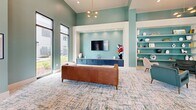 Located at 185 Marquis Way in Freeport, Florida, this single-floor community lounge is a thoughtfully designed social space that blends modern aesthetics with relaxed, inviting comfort. The room is defined by its soaring ceilings, which lend a generous sense of openness and volume well beyond what the footprint alone might suggest.

The walls are painted in a soothing sage green, creating a cohesive and calming atmosphere throughout. Tall, black-framed floor-to-ceiling windows line one side of the room, flooding the interior with natural light and offering views of the surrounding landscape. A modern multi-bulb pendant chandelier adds a warm, stylish focal point overhead.

The entertainment area features a built-in media niche with a wall-mounted flat-screen television and a sleek navy-blue credenza below. Adjacent to it, a recessed display alcove with matching teal shelving showcases an array of decorative objects, adding character and visual depth to the space.

Seating is varied and plentiful, including a tufted cognac leather sofa, plush velvet armchairs in muted tones, and soft ottomans arranged over a vibrant, multi-toned area rug. A full-size pool table and foosball table occupy the central and peripheral areas, making this an ideal space for recreation and social gatherings. The overall ambiance is modern, welcoming, and well-appointed.