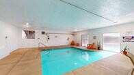 This amenity space serves as a comprehensive recreational facility set within a multi-story residential building complex. At its heart is a generously sized indoor swimming pool, enclosed within a bright, white-painted brick room with a textured ceiling. The pool area is thoughtfully equipped with safety equipment, entry handrails, and casual seating in the form of classic Adirondack chairs. A glazed door allows natural light to filter in, offering views of the outdoor grounds. Adjacent to the pool, a dedicated fitness area is visible through a large pass-through window, housing cardio and strength training equipment. A seating area with a table and chairs provides a comfortable spot to relax nearby. Outside, the grounds open up into a well-maintained courtyard shared between two multi-story brick towers. The outdoor area features a swimming pool surrounded by a spacious patio with lounge chairs, a hammock stand, and umbrella-shaded dining furniture. Lush green lawns, mature trees, ornamental shrubs, and stone-edged garden beds create a serene, park-like atmosphere. Covered carport parking is accessible from the grounds, and iron fencing defines the boundaries of this private outdoor retreat.