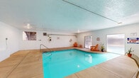 This amenity space serves as a comprehensive recreational facility set within a multi-story residential building complex. At its heart is a generously sized indoor swimming pool, enclosed within a bright, white-painted brick room with a textured ceiling. The pool area is thoughtfully equipped with safety equipment, entry handrails, and casual seating in the form of classic Adirondack chairs. A glazed door allows natural light to filter in, offering views of the outdoor grounds. Adjacent to the pool, a dedicated fitness area is visible through a large pass-through window, housing cardio and strength training equipment. A seating area with a table and chairs provides a comfortable spot to relax nearby. Outside, the grounds open up into a well-maintained courtyard shared between two multi-story brick towers. The outdoor area features a swimming pool surrounded by a spacious patio with lounge chairs, a hammock stand, and umbrella-shaded dining furniture. Lush green lawns, mature trees, ornamental shrubs, and stone-edged garden beds create a serene, park-like atmosphere. Covered carport parking is accessible from the grounds, and iron fencing defines the boundaries of this private outdoor retreat.