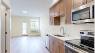 This single-floor unit offers a thoughtfully designed layout that balances functionality with modern aesthetics. The open-concept kitchen flows seamlessly into the living area, creating a bright and connected living space. The kitchen is outfitted with warm-toned wood cabinetry — featuring both upper and lower storage — paired with sleek white quartz countertops and stainless steel appliances, including a range, over-the-range microwave, dishwasher, and a deep double-basin sink with a brushed nickel faucet. Light hardwood-style flooring extends throughout the unit, lending a sense of warmth and continuity across all areas. The living area is filled with natural light, courtesy of a large window and a glass-panel door that opens to a balcony, offering pleasant outdoor views. A flush ceiling light fixture adds supplemental illumination. The bathroom continues the cohesive design language, featuring matching wood cabinetry with a white countertop vanity, light flooring, and clean white walls. The bedroom is a calm, versatile space with the same light flooring and neutral walls, providing a blank canvas for personalization. Overall, the unit presents a clean, contemporary finish throughout.