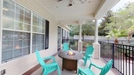 Located at 2220 Gloria Cir in Pensacola, Florida, this single-story home presents a charming and inviting exterior defined by classic Southern architectural details. The standout feature of this property is its spacious covered front porch, which showcases elegant white columns, a tongue-and-groove ceiling, and a ceiling fan — offering both comfort and style in the Florida climate. The porch is designed for outdoor living, centered around a round fire pit table surrounded by vibrant teal Adirondack chairs, creating an ideal gathering space for relaxed conversation and evening enjoyment. Black wrought iron railings frame the perimeter, adding a refined touch while maintaining open views of the lush, tree-lined backyard beyond the white privacy fence. The home's exterior features light-colored siding accented by dark shutters, contributing to a clean, classic aesthetic. Large windows with horizontal blinds allow for natural light throughout the interior while maintaining privacy. The mature trees visible from the porch provide a sense of seclusion and natural beauty, enhancing the overall ambiance of this well-appointed Pensacola residence.