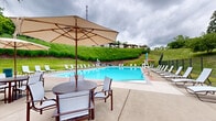 Located at 651 Oaklynn Court in Pittsburgh, PA, this outdoor recreational facility centers around a generously sized swimming pool with vivid blue water and a clearly marked shallow entry area featuring tiered steps for easy access. The pool is surrounded by a wide concrete deck outfitted with rows of white lounge chairs, providing ample space for sunbathing and relaxation. Umbrella-shaded bistro tables and chairs are positioned poolside, offering a comfortable setting for casual gathering. A dedicated single-story clubhouse structure anchors one end of the facility, featuring an outdoor patio covered by multiple large umbrellas and furnished with dining sets, ideal for socializing. The clubhouse appears to include restroom and support facilities for pool guests. The entire pool area is enclosed by a black metal fence, ensuring a secure environment. The surrounding landscape is lush and well-maintained, with rolling green hillsides, ornamental grasses, and mature trees framing the space and providing a natural, park-like backdrop. The open-air setting takes full advantage of the outdoor environment, offering views of the verdant surroundings. Overall, the facility presents a well-appointed, community-oriented outdoor amenity space suited for warm-weather recreation.