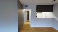 This single-floor space offers a practical and modern layout designed for comfortable everyday living. The interior is finished with warm-toned hardwood flooring that flows seamlessly throughout the open living area and hallway, adding a sense of continuity and warmth to the space.

The kitchen is positioned adjacent to the main living area and features a raised countertop that serves as a natural divider between the two spaces. Dark upper cabinetry contrasts with light-colored lower cabinets and countertops, creating a striking visual balance. Under-cabinet lighting illuminates the workspace, enhancing both functionality and ambiance.

The walls are finished in a cool, muted gray tone that complements the flooring and cabinetry, lending the space a clean, contemporary feel. White baseboards and door trim provide crisp contrast throughout. A central hallway connects the living area to the remaining rooms, with a doorway visible at the end leading to additional spaces. The textured ceiling adds a subtle architectural detail, while a centrally located HVAC vent ensures efficient climate control. Overall, the space presents a well-organized, move-in-ready environment with a cohesive modern aesthetic.