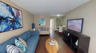 This single-story unit offers a thoughtfully organized layout with one bedroom and one bathroom. The living area serves as the focal point of the space, featuring dark hardwood-style flooring that extends throughout the main areas, warm neutral-toned walls, and a textured ceiling. The open arrangement allows the living room to flow naturally into a dedicated dining area, making the overall space feel connected and functional. The living room provides ample room for seating and entertainment, with a media console positioned opposite the main seating area.

The bedroom is carpeted for added comfort and is equipped with a ceiling fan with integrated lighting. A closet with shelving is accessible directly from the bedroom, offering organized storage. A large window with vertical blinds allows natural light to fill the room.

The bathroom features a full tub and shower combination with a curtain enclosure and white tile surround. Clean white fixtures, including a toilet and vanity, complete the space with a straightforward, functional design.

Throughout the unit, neutral wall tones create a cohesive backdrop, while the consistent flooring choices between areas reinforce a unified aesthetic. The overall layout is efficient, making practical use of the available space.
