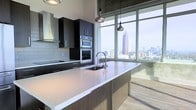 This contemporary high-rise residence offers a refined urban living experience highlighted by breathtaking panoramic city skyline views. The open-concept kitchen serves as the centerpiece of the space, showcasing a sophisticated design with dark espresso cabinetry that contrasts elegantly against light-toned quartz countertops. A large kitchen island with an undermount sink and modern pendant lighting provides both functional workspace and a stylish focal point. Stainless steel appliances, including a double wall oven and a sleek range hood, underscore the kitchen's professional-grade quality. Floor-to-ceiling windows flood the interior with abundant natural light while framing a dramatic cityscape, with the distinctive red-topped Bank of America Plaza tower — an Atlanta landmark — prominently visible on the horizon. The light wood flooring adds warmth and continuity throughout the space, complementing the modern aesthetic. Exposed ceiling elements lend an industrial edge to the overall design. The single-floor layout is thoughtfully designed to maximize the sense of openness and connection to the vibrant urban environment outside, making this residence an ideal setting for those who appreciate contemporary design and city living.