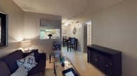 This one-bedroom, one-bathroom unit is thoughtfully laid out across a single floor, maximizing functionality within a compact footprint. The living room serves as the central gathering space, featuring neutral-toned walls, textured ceilings, and carpeted flooring. A large wall mirror and ambient lamp lighting contribute to an open, welcoming feel. The room connects seamlessly to a dining area with a ceiling fan overhead and a pass-through window into the kitchen, enhancing the sense of flow between spaces.

The galley-style kitchen is efficiently organized with wood-toned cabinetry, granite-style countertops on both sides, a stainless steel sink, and standard appliances including an electric range and full-size refrigerator. Wood-look flooring adds warmth to this functional space.

The bedroom is a comfortable retreat, featuring plush carpeting, a window with blinds and floor-length curtains allowing natural light control, and wall art above the bed. The room offers a calm, restful atmosphere with warm neutral tones throughout.

The bathroom includes a full tub-shower combination with a curtain, a granite-style vanity countertop, a large mirror, and wood-look flooring, providing a clean and practical design. Overall, the unit presents a cohesive, cozy aesthetic suited for efficient single-floor living.