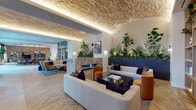 This thoughtfully designed amenity lounge offers a refined and welcoming environment distinguished by its striking architectural details and curated interiors. The space is anchored by a dramatic textured ceiling with warm integrated uplighting that creates an inviting, golden ambiance throughout. Large-format tile flooring transitions seamlessly into herringbone wood-patterned flooring, defining distinct zones across the open layout.

A standout living wall installation—featuring lush ferns, trailing plants, and upright greenery set against a dark stone planter—serves as a natural focal point along one wall, complemented by perforated wall panels that add textural depth. Multiple seating vignettes are thoughtfully arranged throughout, including plush sofas, accent armchairs in warm amber and cobalt blue tones, and a communal wooden conference table with upholstered seating—ideal for both casual lounging and collaborative work.

A double-height window nook provides an intimate retreat flooded with natural light and framing views of an adjacent outdoor terrace and surrounding urban skyline. Steel-framed glass partition walls delineate areas while maintaining openness and visual connectivity. Built-in shelving with curated décor, an open display unit, and artful accessories complete the space, offering both function and aesthetic charm throughout this elegantly layered interior.