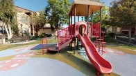 This outdoor recreational space is designed to serve as a community playground within a well-maintained residential setting. The centerpiece is a multi-level play structure featuring bold red railings, a covered upper platform, multiple access points including a ramp and climbing ladder, and a curved red slide. The structure is built with safety-conscious design, incorporating perforated metal decking and enclosed side panels with window cutouts on the platform level.

The play area sits atop a colorful rubber safety surface featuring soft, organic shapes in muted tones of blue, green, pink, and tan, providing cushioned ground cover throughout the active zone. This transitions into a paved pathway that winds through the surrounding green lawn, where two wooden park benches offer comfortable seating for supervising adults or visitors.

The space is framed by mature deciduous trees that provide partial shade and a natural canopy overhead. Well-kept shrubs and manicured landscaping line the perimeter, contributing to the overall tidiness of the environment. The surrounding multi-story buildings, finished in warm beige stucco with tile roofing, are visible in the background, reinforcing the community-oriented character of the space. The area benefits from ample natural sunlight throughout the day.