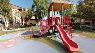 This outdoor recreational space is designed to serve as a community playground within a well-maintained residential setting. The centerpiece is a multi-level play structure featuring bold red railings, a covered upper platform, multiple access points including a ramp and climbing ladder, and a curved red slide. The structure is built with safety-conscious design, incorporating perforated metal decking and enclosed side panels with window cutouts on the platform level.

The play area sits atop a colorful rubber safety surface featuring soft, organic shapes in muted tones of blue, green, pink, and tan, providing cushioned ground cover throughout the active zone. This transitions into a paved pathway that winds through the surrounding green lawn, where two wooden park benches offer comfortable seating for supervising adults or visitors.

The space is framed by mature deciduous trees that provide partial shade and a natural canopy overhead. Well-kept shrubs and manicured landscaping line the perimeter, contributing to the overall tidiness of the environment. The surrounding multi-story buildings, finished in warm beige stucco with tile roofing, are visible in the background, reinforcing the community-oriented character of the space. The area benefits from ample natural sunlight throughout the day.