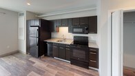 This single-floor unit presents a cohesive modern design throughout its open layout. The kitchen is a standout feature, equipped with dark espresso cabinetry extending from floor to ceiling, light granite-style countertops, and a full suite of black appliances including a top-freezer refrigerator, electric range with overhead microwave, and dishwasher. Under-cabinet lighting and an exposed overhead duct add both functionality and an industrial design element. Wood-look plank flooring runs continuously through the kitchen and into the open living area, creating a seamless visual flow. The living space is generously sized, featuring neutral white walls, recessed lighting, and a large sliding glass door that opens to a private balcony, allowing ample natural light to fill the room. A hallway connects the living area to additional rooms, including a closet for added storage. The bathroom maintains the same design language, featuring a dark vanity with granite-style countertops, wood-look flooring, brushed nickel fixtures, and a full soaking tub with an overhead shower. Grab bars and clean white walls complete the bathroom's functional and polished aesthetic.