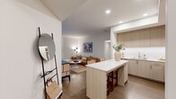 This thoughtfully designed single-floor residence offers a seamless blend of modern aesthetics and functional living. The open-concept layout connects the kitchen and living area, creating an inviting and cohesive space ideal for both relaxing and entertaining. The kitchen is a standout feature, outfitted with flat-front light wood cabinetry extending to the ceiling, a white quartz waterfall island with seating for two, a white tile backsplash, and a full suite of stainless steel appliances. Light hardwood flooring flows throughout the main living areas, complementing the warm, neutral tones of the space. The living room is anchored by a tan leather sectional sofa, a round white coffee table, and a stylish floor lamp, with sliding glass doors providing direct access to an outdoor area and allowing natural light to fill the room. The primary bedroom is bright and airy, featuring large floor-to-ceiling sliding doors that open to the outdoors. A hallway connects the bedroom to additional rooms, including a secondary bathroom with a soaking tub and shower combination. The primary bathroom features large-format cream tile flooring, a wide wood-toned vanity with a white countertop, and a backlit mirror. Throughout, the clean white walls and recessed lighting create a calm, sophisticated ambiance.